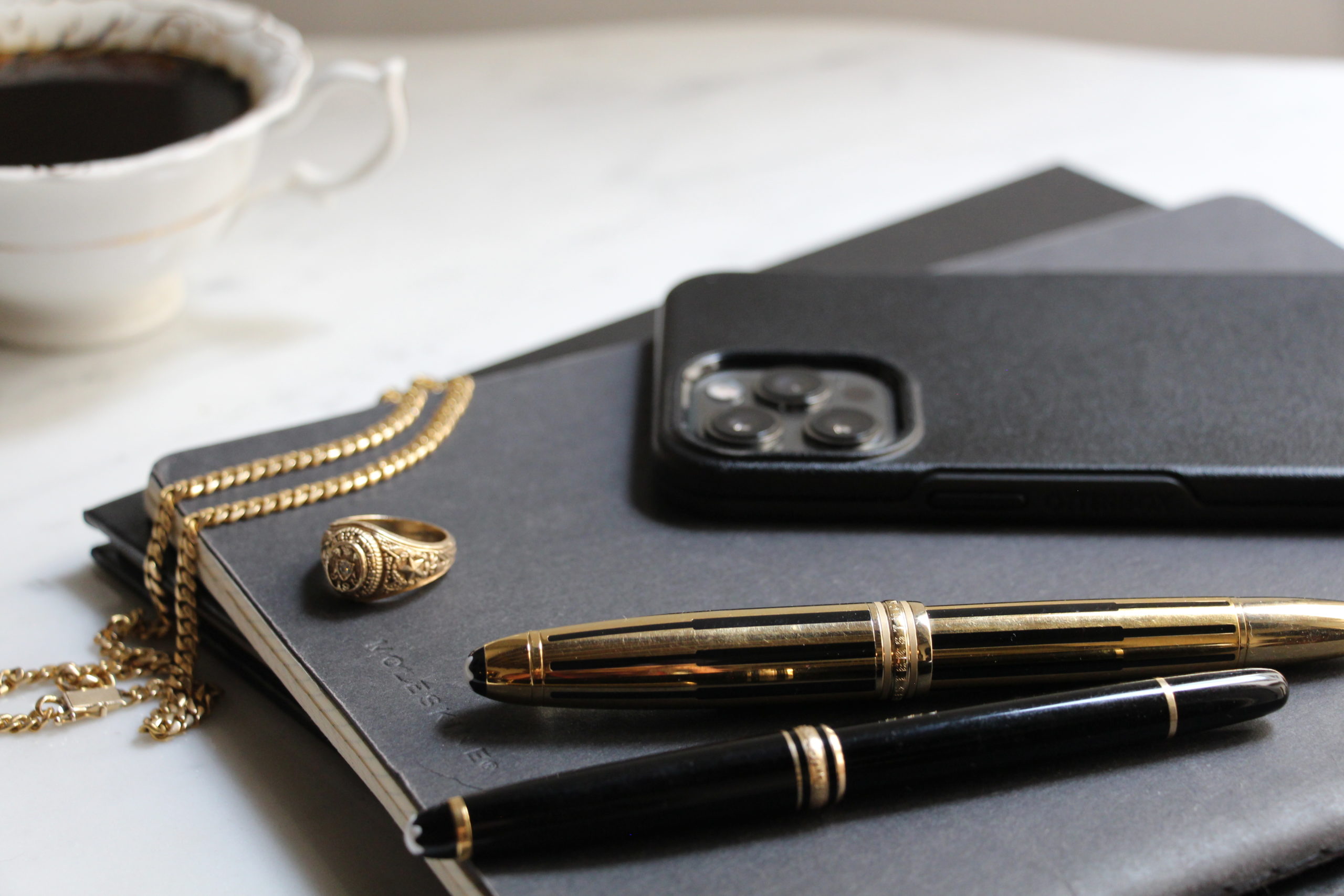 iPhone 12 Pro Max black case with Mont Blanc pens, a coffee cup, gold necklace and gold ring